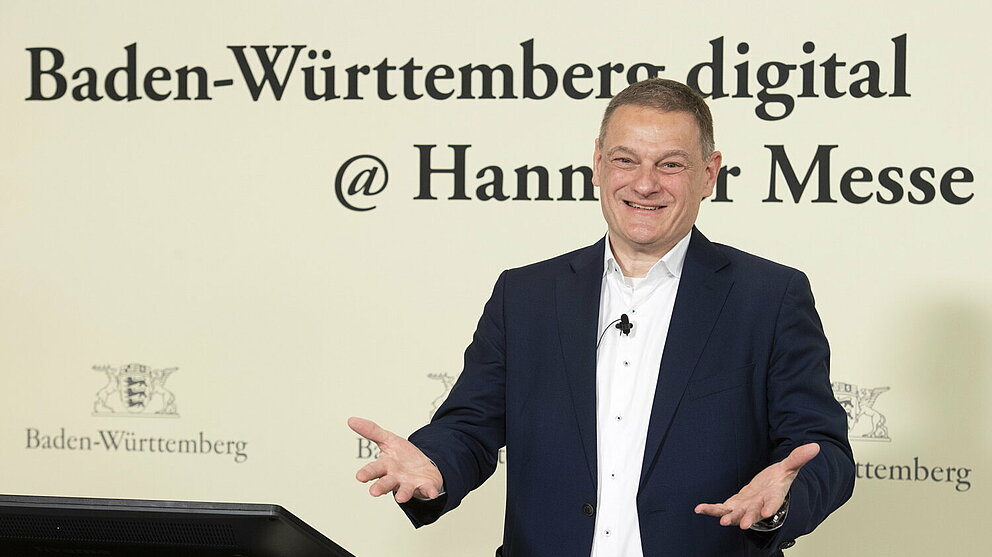 Franz Loogen blickt lächelnd in die Kamera. Er steht vor einer Wand mit dem Schriftzug 'Baden-Württemberg digital @ Hannover Messe'. Seine Hände sind leicht nach vorne ausgestreckt, um die Gäste zu begrüßen.. 