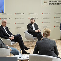 Die Podiumsteilnehmenden am Zulieferertag Automobilwirtschaft BW 2020 sitzen im Halbkreis, im Hintergrund ist ein Referent per Video zugeschaltet.