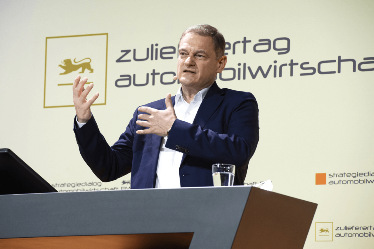 Franz Loogen begrüßt die Teilnehmenden zum Zulieferertag Automobilwirtschaft BW, er steht vor einem Rednerpult und hat die Hände gehoben.