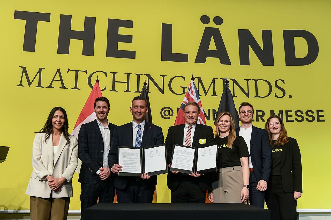 Sieben Personen blicken in die Kamera. Zwei halten einen Vertrag in der Hand. Im Hintergrund sieht man eine gelbe Messewand von THE LÄND.