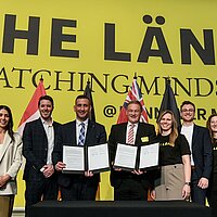 Sieben Personen blicken in die Kamera. Zwei halten einen Vertrag in der Hand. Im Hintergrund sieht man eine gelbe Messewand von THE LÄND.