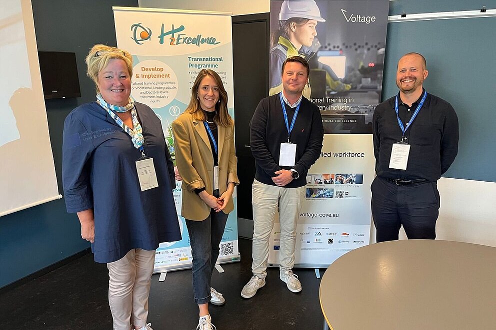 Four people stand in a room during the workshop at the Forum on Vocational Excellence in Kolding, Denmark. On the left a banner of the H2Excellence project and on the right a banner of the Voltage project.