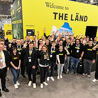 Gruppenfoto von Studierenden am Baden-Württemberg Pavillon auf der Hannover Messe. Die Studentinnen und Studenten winken in die Kamera.