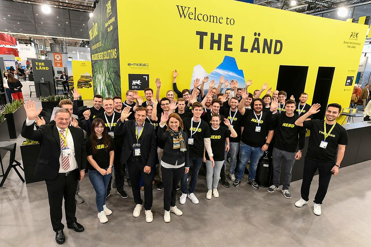 Gruppenfoto von Studierenden am Baden-Württemberg Pavillon auf der Hannover Messe. Die Studentinnen und Studenten winken in die Kamera.