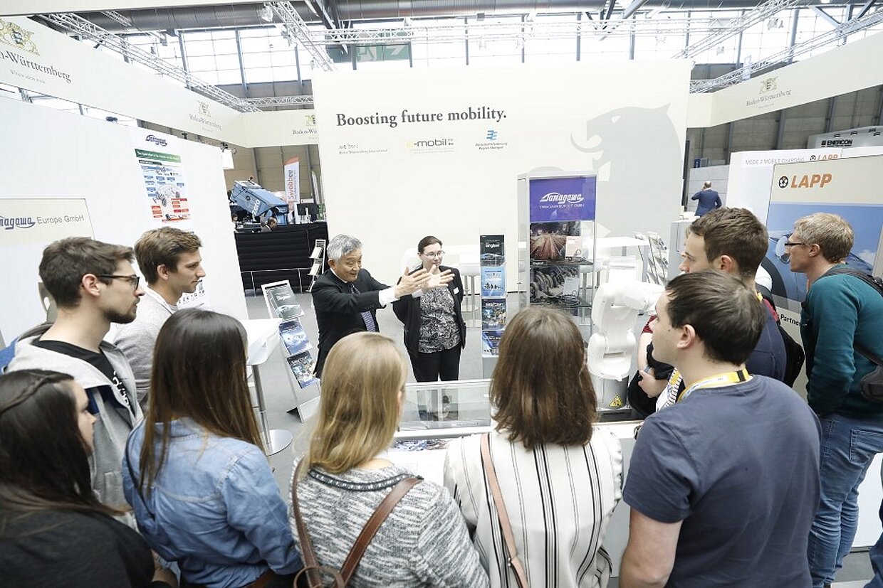 Hannnover Messe Study Tours Studenten an einem Stand auf der Hannover Messe erhalten einen Vortrag von einem Mann in einem Anzug.