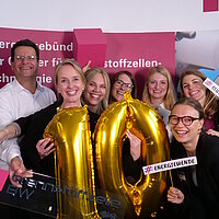 Gruppenfoto des Teams Wasserstofftechnologien der e-mobil BW, einige halten Schilder in der Hand mit der Aufschrift "#Wasserstoff" und die Person im Vordergrund hält eine "10" aus zwei goldenen Ballons in der Hand.