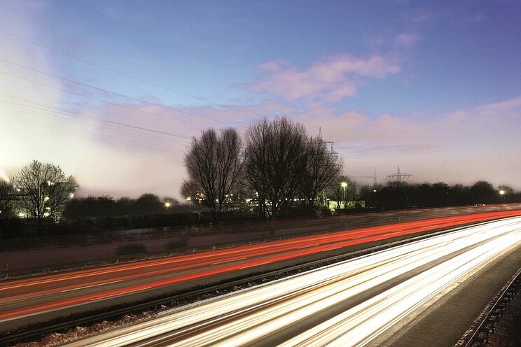 Abendaufnahme einer Autobahn bei Sonnenuntergang. Der Himmel ist in violett-lila Tönen gefärbt. Die Autobahn selbst ist nur angedeutet sichtbar durch leuchtende Lichtschlieren der fahrenden Autos