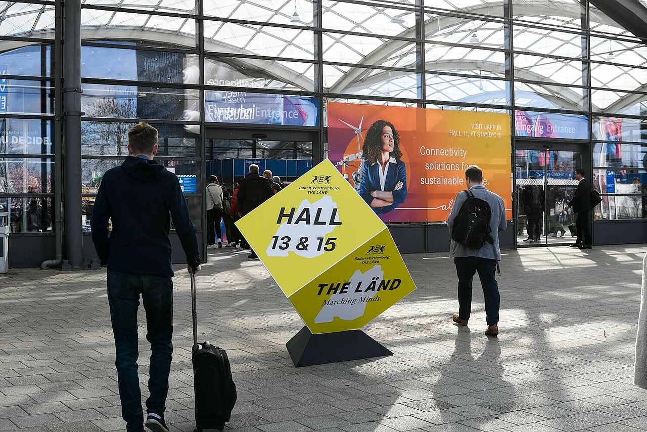 Außenaufnahme auf der Hannover Messe. Ein gelber Würfel in den Baden-Württemberg-Farben weist auf den THE LÄND-Pavillon hin.