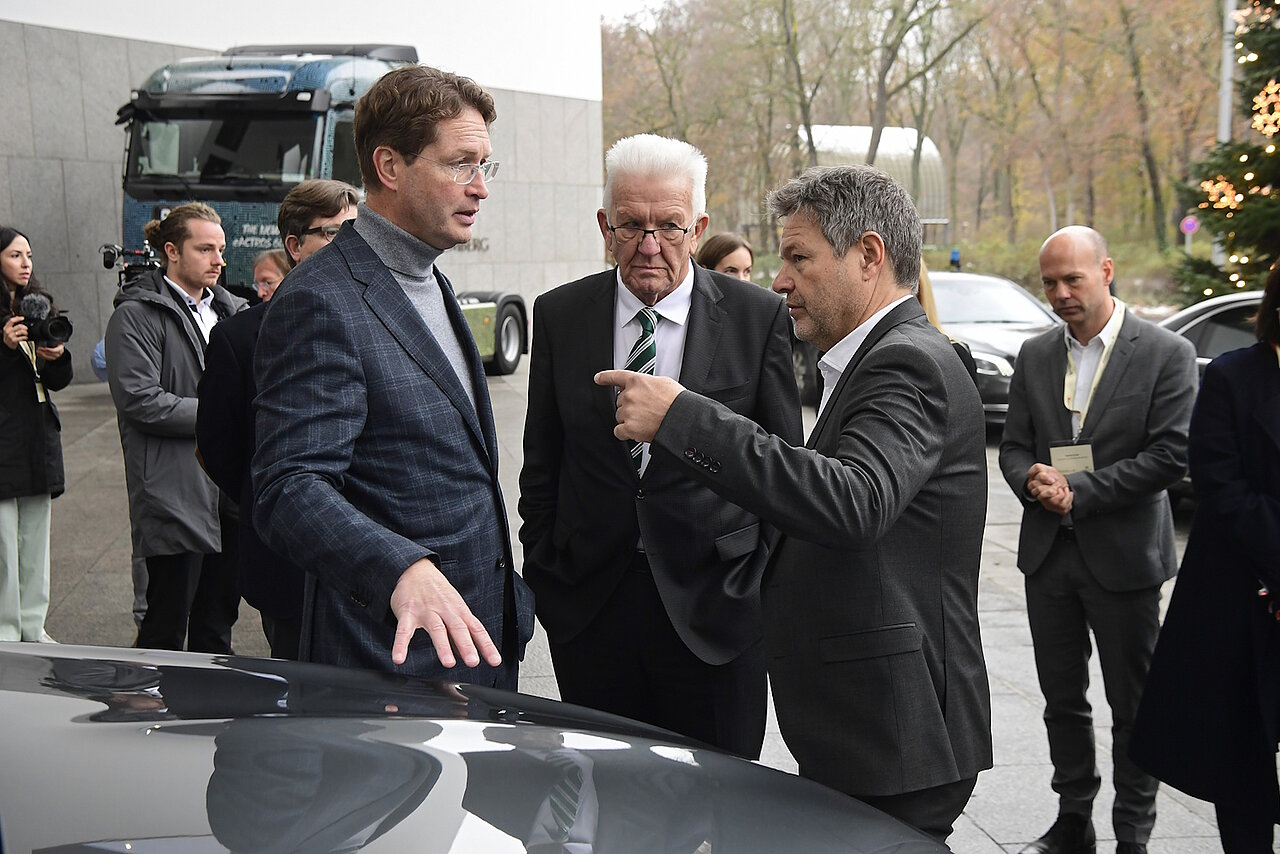 Baden-Württembergs Ministerpräsident Winfried Kretschmann steht mit zwei Personen vor einem Mercedes Pkw und unterhält sich.