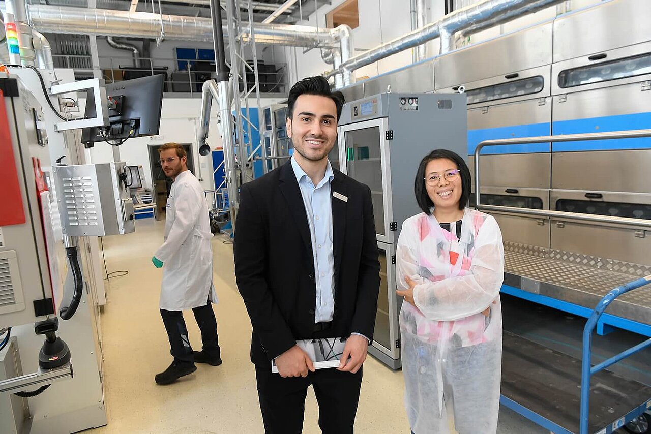 Zwei Personen blicken lächelnd in die Kamera. Sie stehen in einer Forschungshalle für Batterietechnologie.