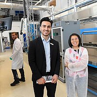 Zwei Personen blicken lächelnd in die Kamera. Sie stehen in einer Forschungshalle für Batterietechnologie.