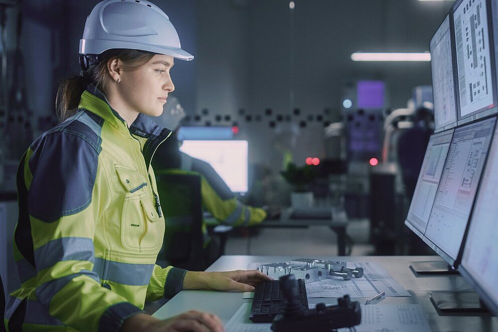 Seitliche Aufnahme einer jungen Frau mit Schutzhelm und Arbeitskleidung. Sie blickt konzentriert auf mehrere große Bildschirme mit technischen Daten.