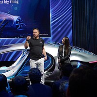 Show eines neuen Konzeptfahrzeug auf der Messe. Im Vordergrund spricht ein Referent lebendig von neuen Design und Funktionen des Autos.