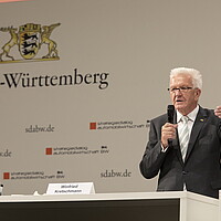 Ministerpräsident Winfried Kretschmann steht auf der Bühne und spricht in ein Mikrofon.