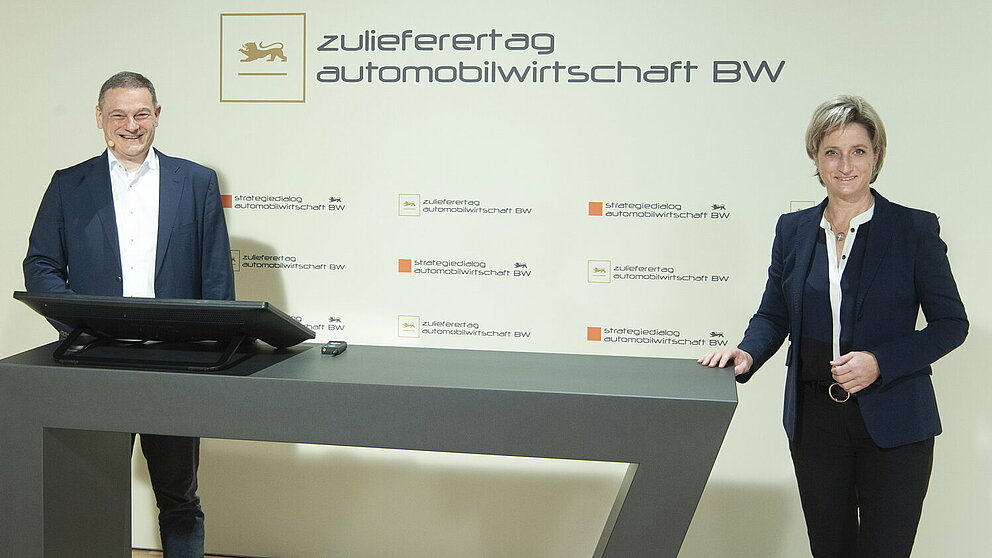 Zwei Personen stehen an einem Sprechpult und blicken lächelnd in die Kamera. Hinter ihnen ist das Logo des Zulieferertags Automobilwirtschaft BW zu sehen.