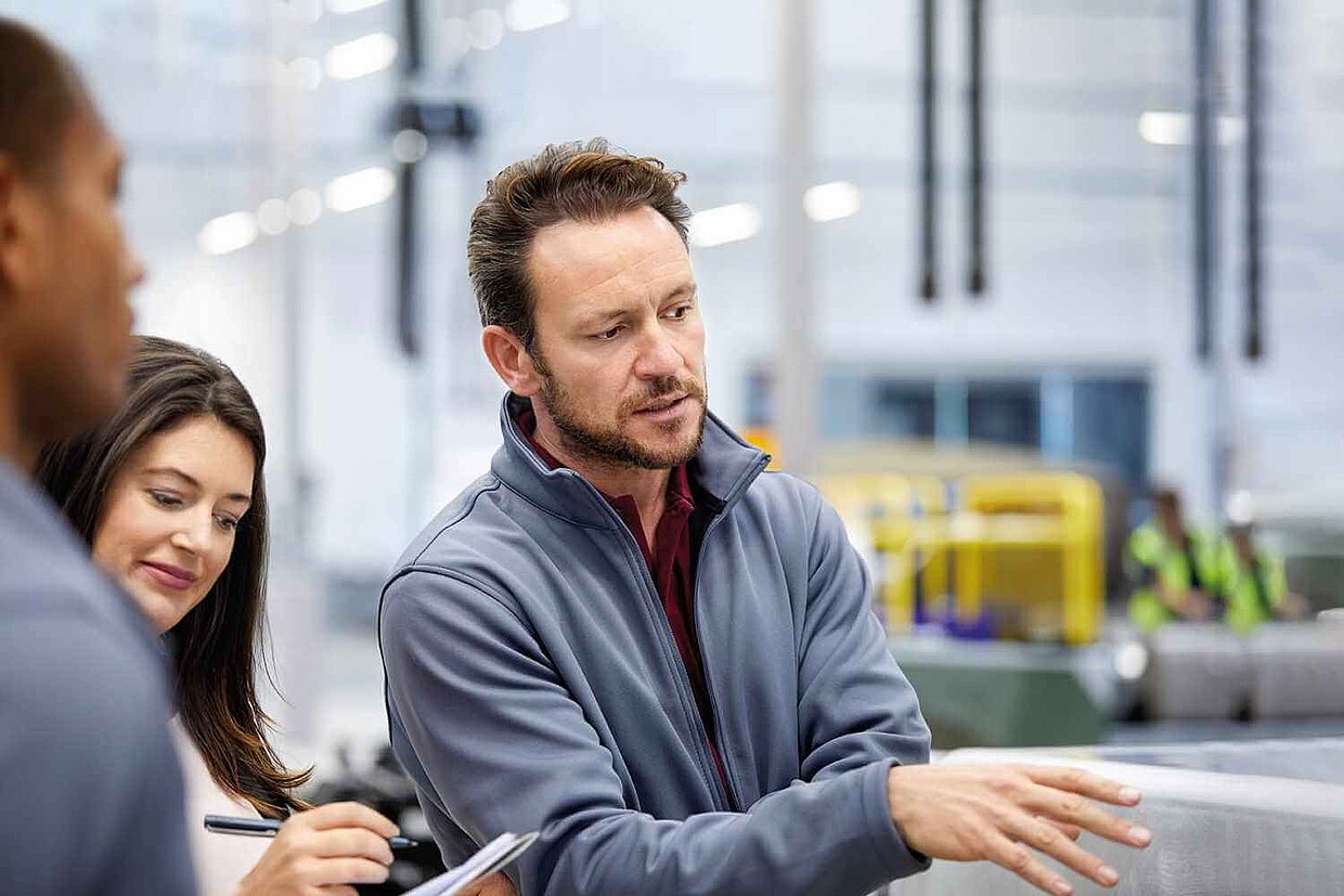 Ein Mann erklärt zwei anderen Personen etwas – alle stehen beisammen und wirken im Gespräch vertieft.