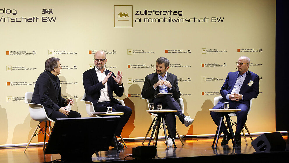Vier Referenten sitzen auf einer Bühne und diskutieren bei einer Podiumsdiskussion, im Hintergrund ist das Logo des Zulieferertags Automobilwirtschaft BW zu sehen.