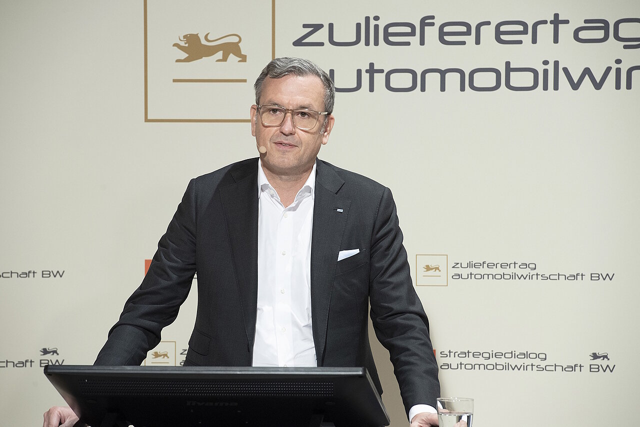 Dr. Jochen Weyrauch steht auf einer Bühne, vor ihm ist ein Monitor und im Hintergrund eine Wand mit der Aufschrift "Strategiedialog Automobilwirtschaft BW"