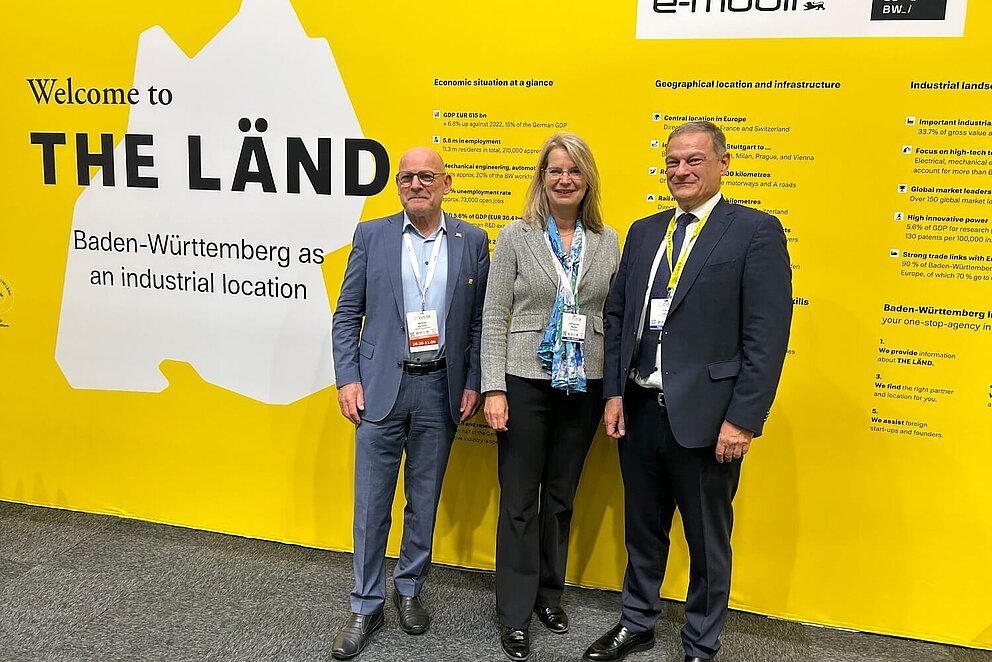 Drei Personen vor einer gelben Messewand auf einer Veranstaltung. Alle Personen blicken in die Kamera.