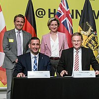Gruppenfoto von vier Personen. Zwei sitzen an einem Tisch und unterschreiben einen Vertrag. Die anderen beiden Personen stehen dahinter. Im Hintergrund sieht man Fahnen und eine gelbe Messewand.