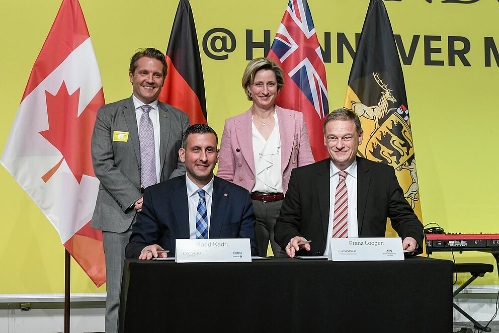 Gruppenfoto von vier Personen. Zwei sitzen an einem Tisch und unterschreiben einen Vertrag. Die anderen beiden Personen stehen dahinter. Im Hintergrund sieht man Fahnen und eine gelbe Messewand.