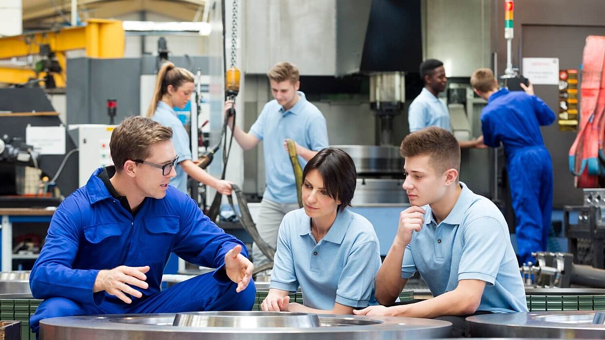 Ein Mann unterrichtet zwei jüngere Personen in einer Produktionshalle. 