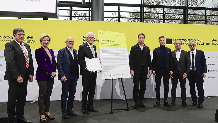 Eine Ansammlung von Personen auf dem Podium des Strategiedialogs Automobilwirtschaft BW. Ministerpräsident Winfried Kretschmann hält ein wichtiges Dokument in der Hand.