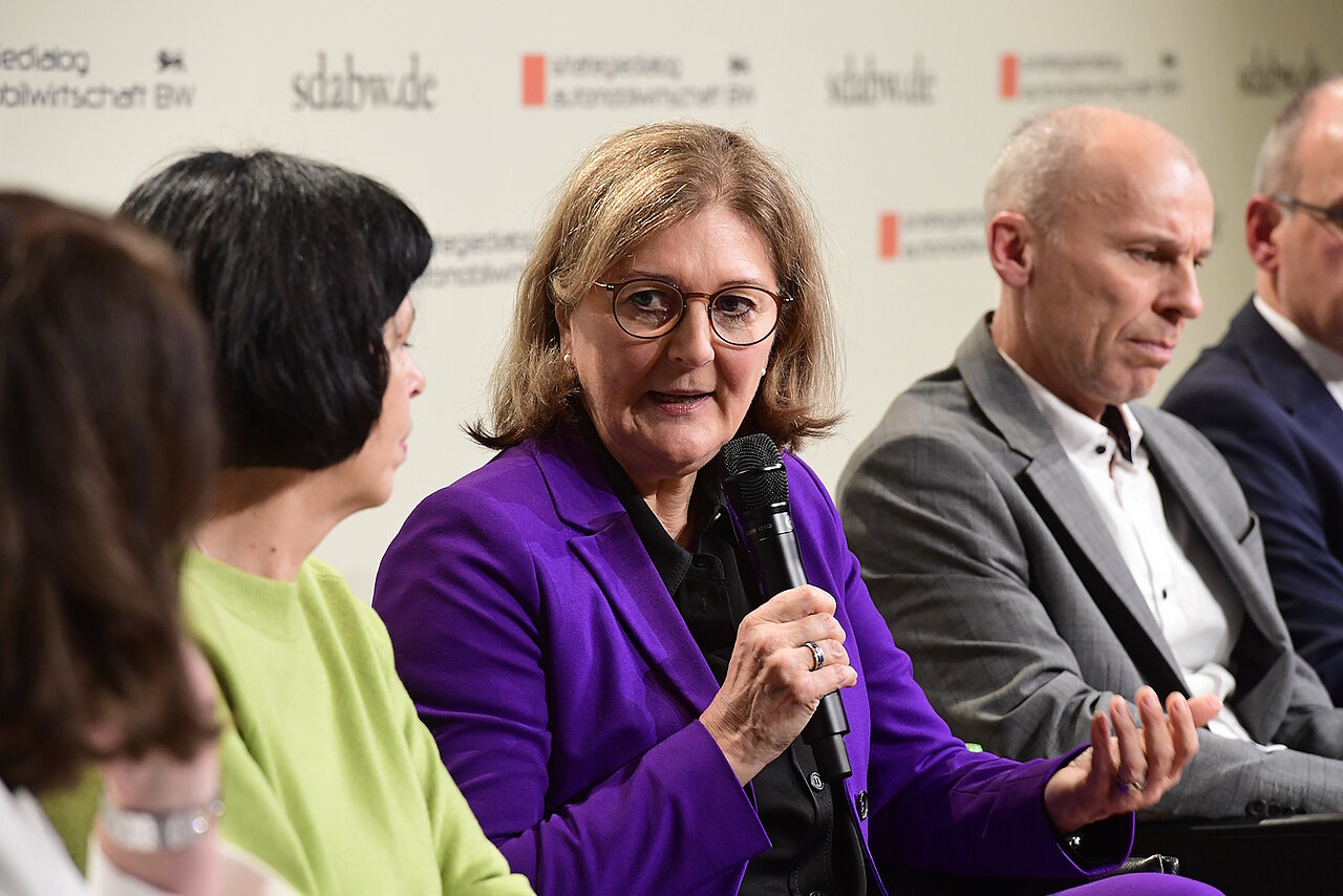 Eine Frau mit einem Mikrofon in der Hand, sie trägt einen lila Blazer.