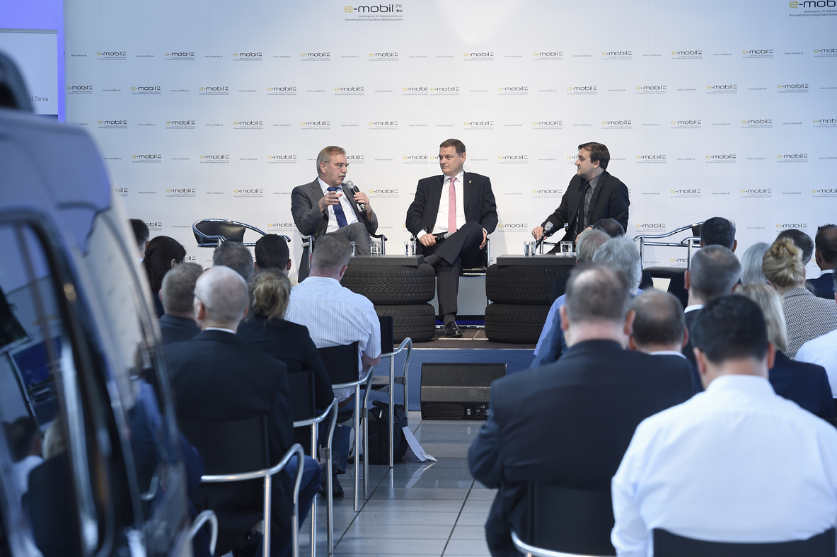 Photo: (c) e-mobil BW Drei Männer im Anzug sitzen auf einer Bühne und führen ein Gespräch. Vor der Bühne sitzt ein Publikum auf Stühlen, das von hinten zu sehen ist.