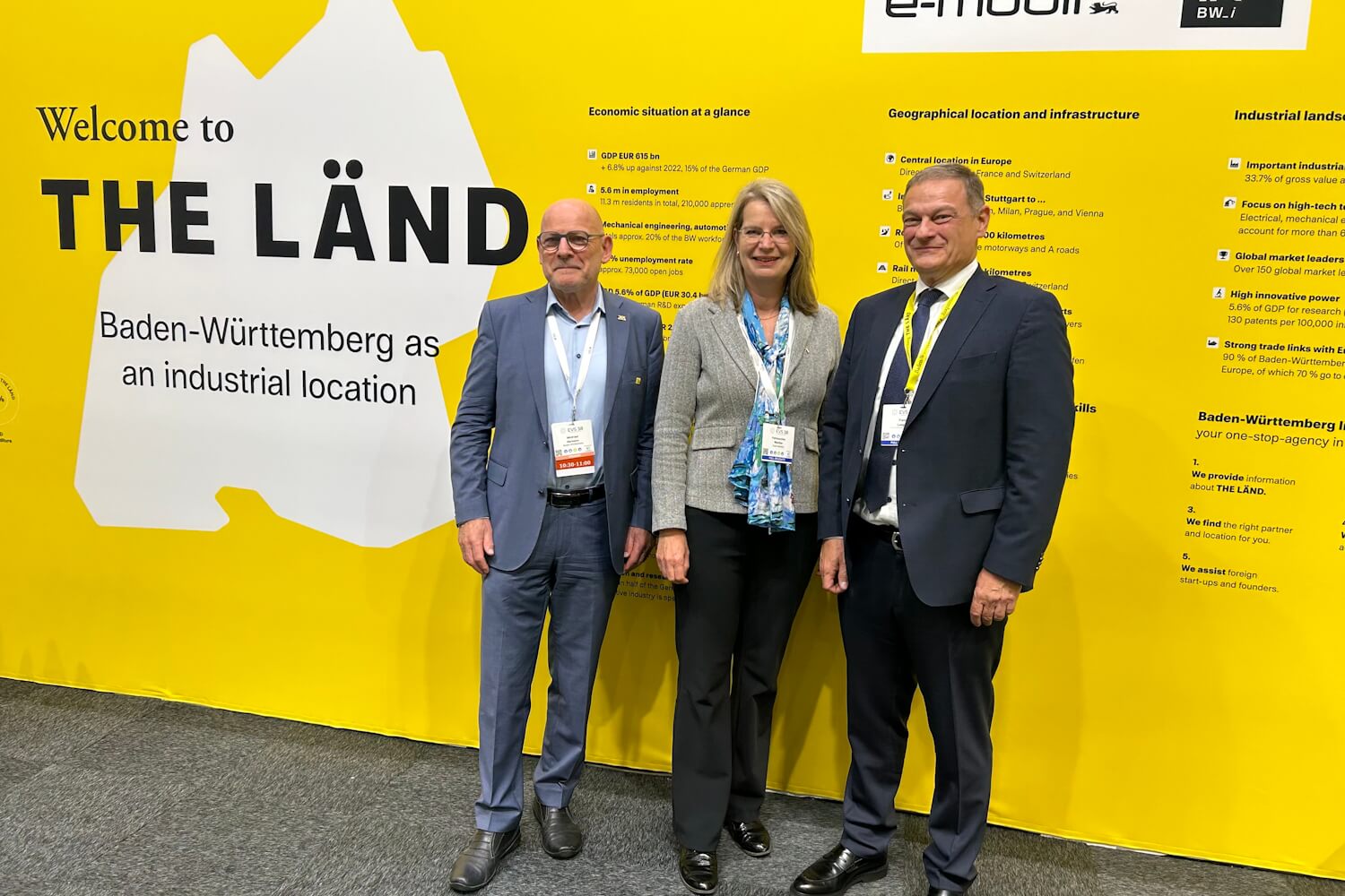 Drei Personen vor einer gelben Messewand auf einer Veranstaltung. Alle Personen blicken in die Kamera.