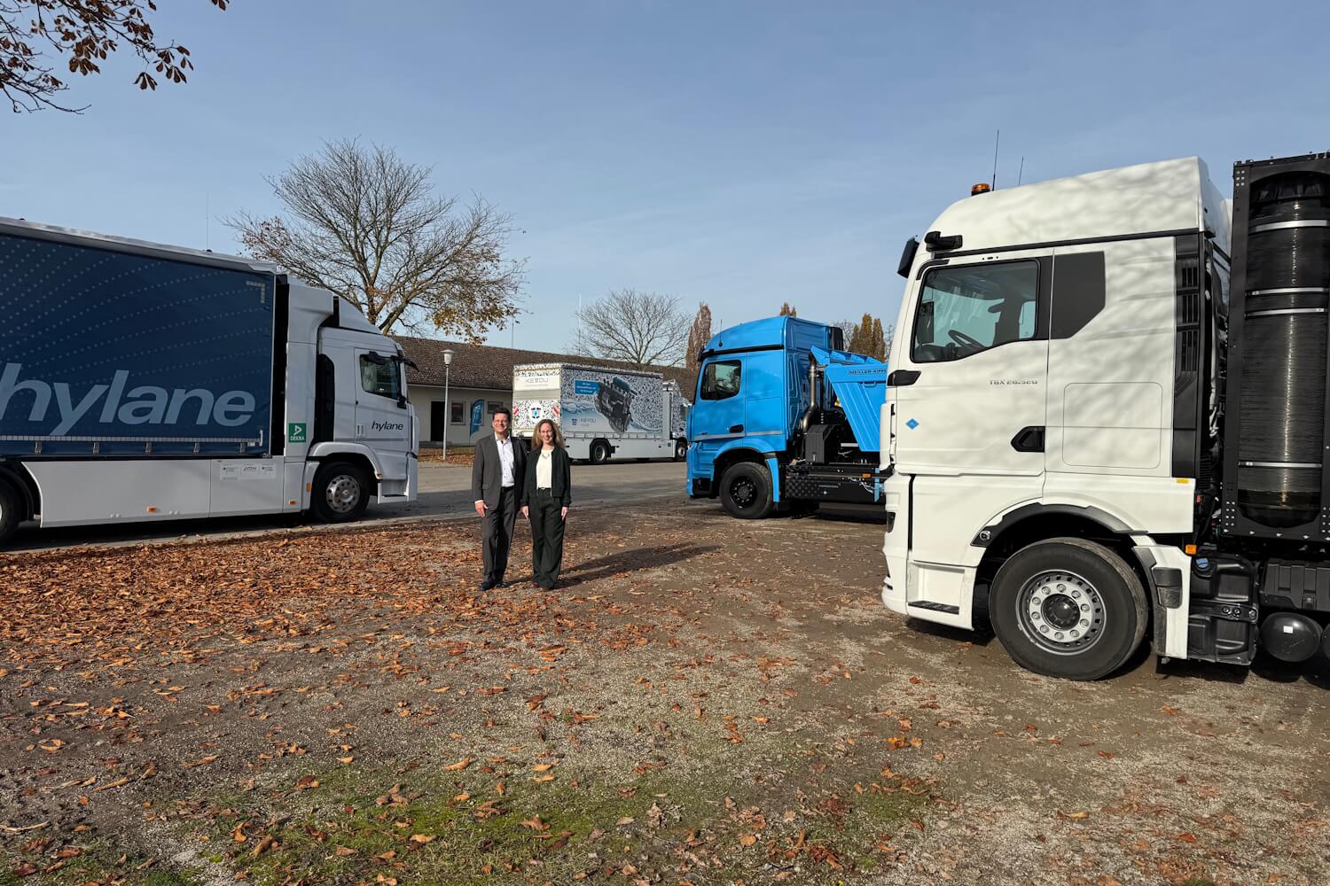 Außenaufnahme von zwei Personen, die in die Kamera lächeln. Im Hintergrund sind Nutzfahrzeuge zu sehen.