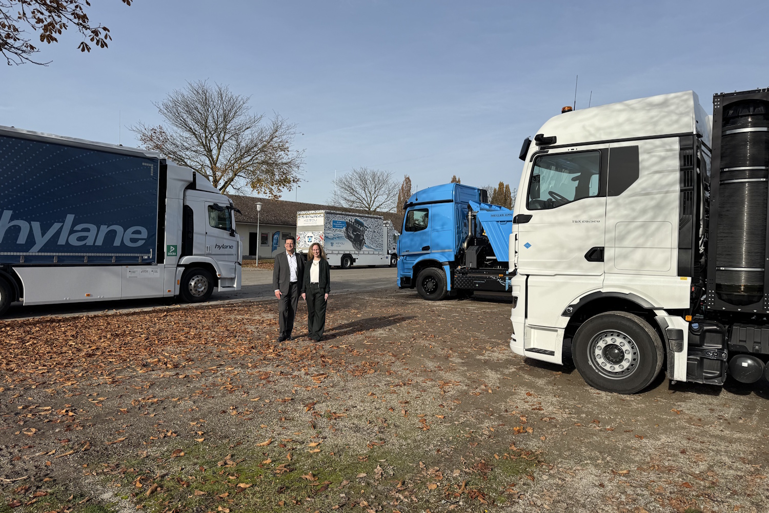 Nutzfahrzeuge und Antrieb mit Wasserstoff Vier Lkw stehen auf einer großen Fläche. In der Mitte stehen zwei Personen, die lächelnd in die Kamera blicken.