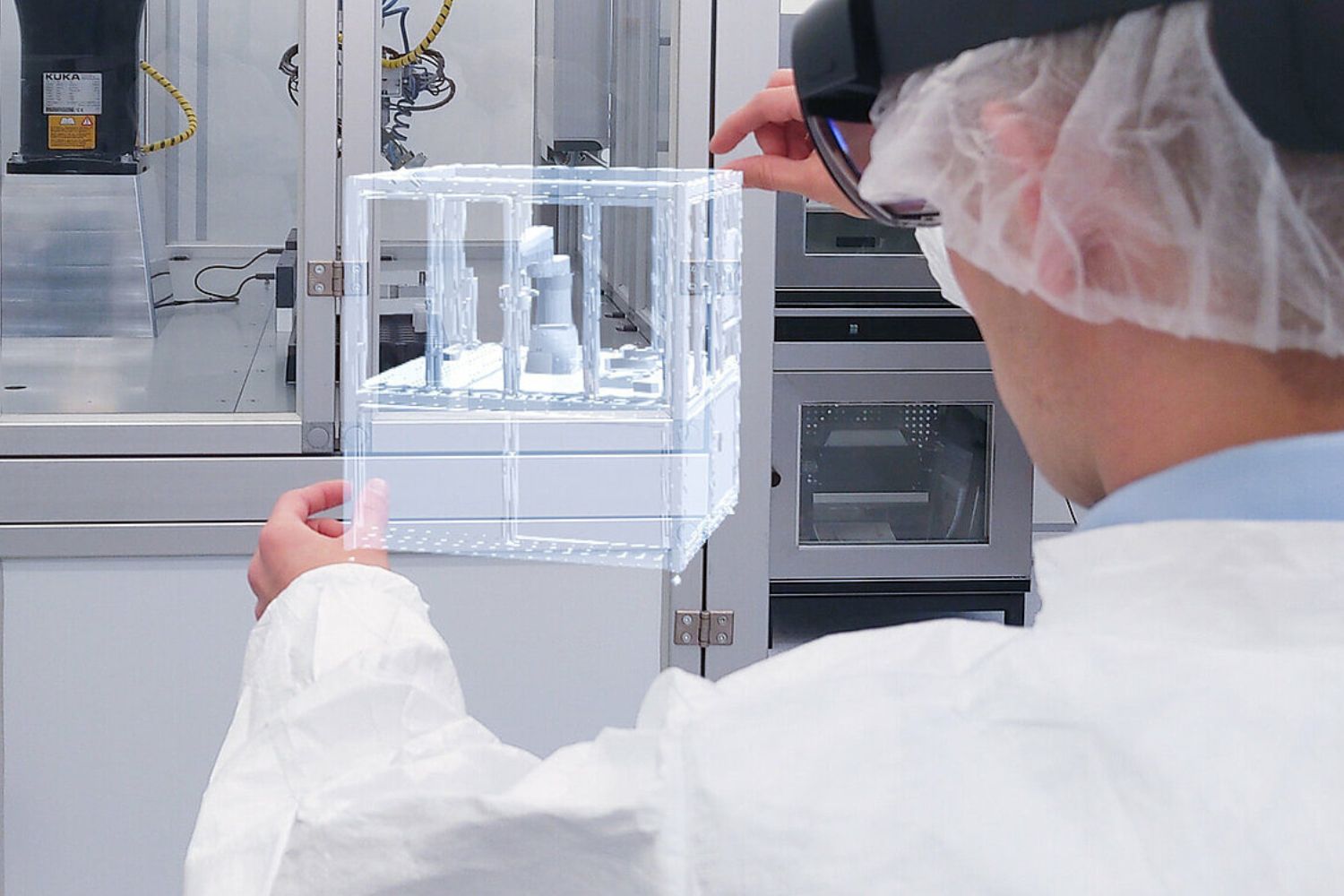 Eine Person in Laborkleidung und Haarnetz betrachtet durch eine Datenbrille eine holografische Darstellung einer Maschine in einem Labor. Die virtuelle Visualisierung zeigt ein technisches Anlagenmodell, das über die reale Umgebung gelegt wird.